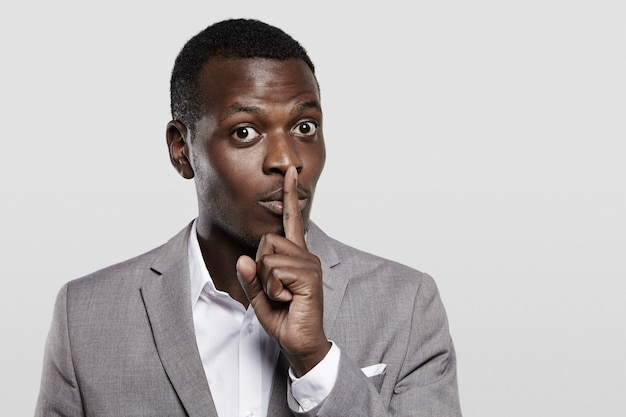 Dark-skinned entrepreneur in gray suit holding finger on his lips, asking to keep confidential information private, concealing commercial secret, saying 'hush'. 