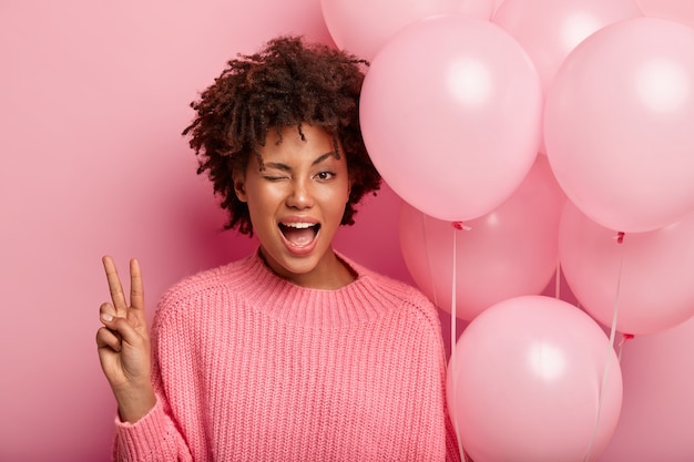 Free photo dark skinned female model blinks eye, shows peace gesture, wears pink jumper and carries air balloons, has fun in circle of best friends, isolated against rosy wall. youth, celebration concept