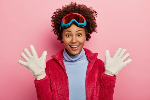 Free photo dark skinned female skier raises palms in white gloves, has fun after snowboarding, laughs joyfully, wears protective mask, turtleneck and red jacket, has winter holidays, isolated on pink background