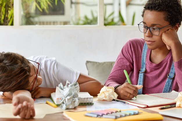 Dark skinned female student wears transparent glasses, looks seriously at tired classmate, work together at course paper, pose at desk with papers and notepad, collaborate to learn material.