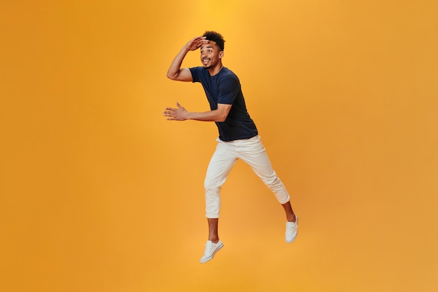 Dark skinned guy in black and white outfit jumping on isolated background Brunette man in tee moving on orange backdrop