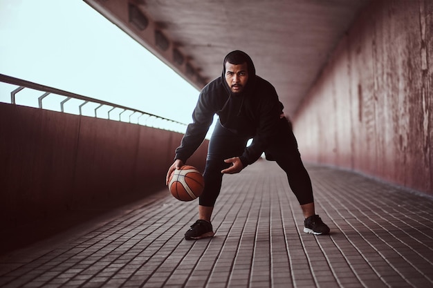 Dark-skinned guy dressed in a black hoodie and sports shorts playing basketball on a footway under a bridge, looking at a camera.