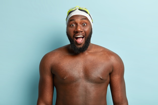 Free photo dark skinned handsome guy with naked body, prepares for swim, wears special hat and goggles, keeps mouth widely opened