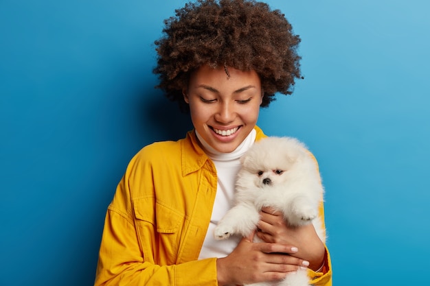 Dark skinned lady keeps lips rounded, wants to kiss adorable pet, plays with small puppy