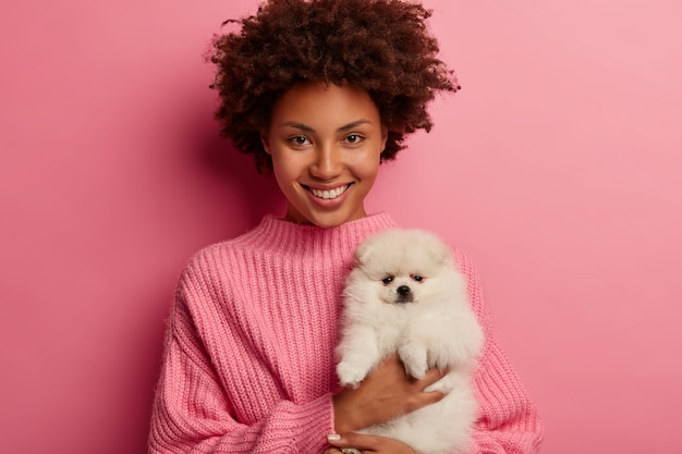 Free photo dark skinned lady keeps lips rounded, wants to kiss adorable pet, plays with small puppy