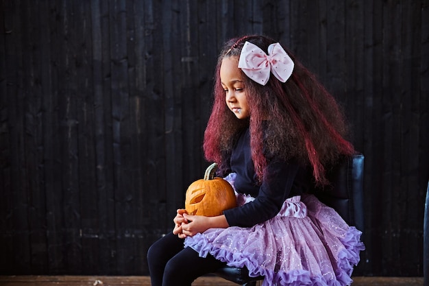 Free photo dark skinned little girl dressed in trendy scary dress holds pumpkin while sitting on chair in dark house. halloween concept.