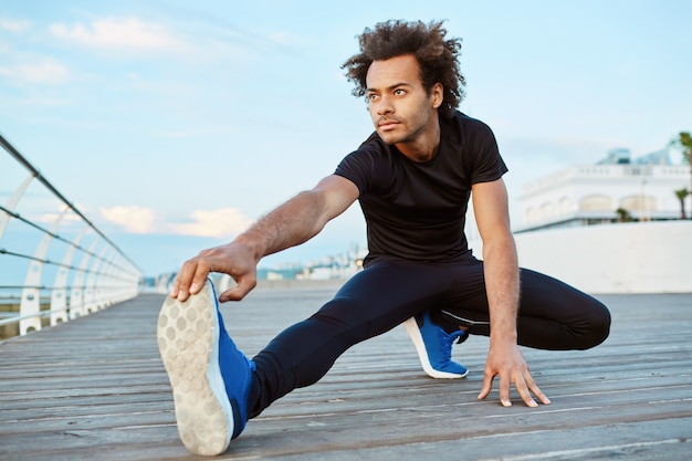 Dark-skinned male athlete with bushy hair doing exercise and stretching legs.