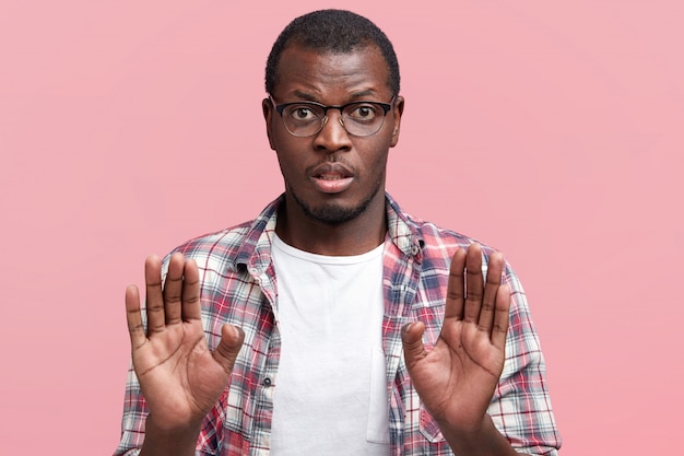 Dark skinned male in eyewear refuses to do something, keeps palms in foreground, denies or rejects to go somewhere, wears spectacles and checkered shirt, isolated over pink