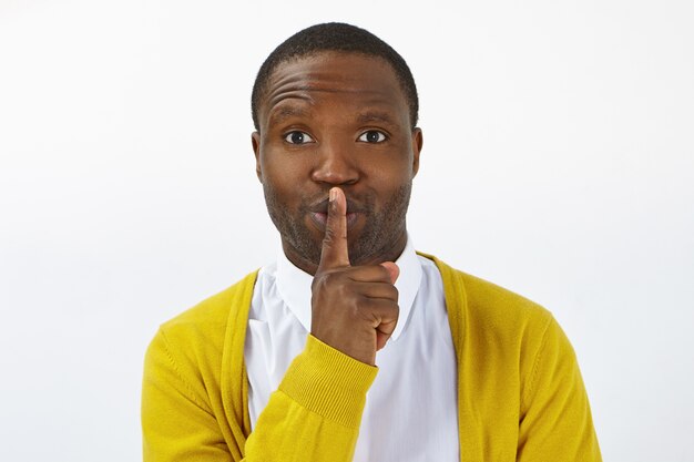 Dark skinned male keeping fore finger on lips, making shush gesture, telling secret or confidential information, asking to be quiet. Handsome young African man shushing in studio, demanding silence