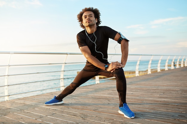 Dark-skinned male runner with beautiful fit body warming-up before cardio workout. Male athlete in sportswear stretching legs with lunge hamstring stretch exercise by the sea in morning sunlight