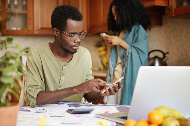 Темнокожий мужчина в очках занимается финансами, используя мобильный телефон, калькулятор и портативный компьютер