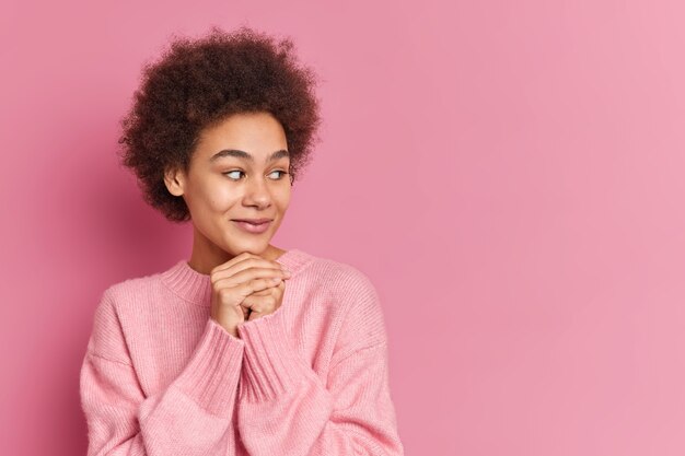 dark skinned millennial girl keeps hands under chin concentrated aside has pleased expression