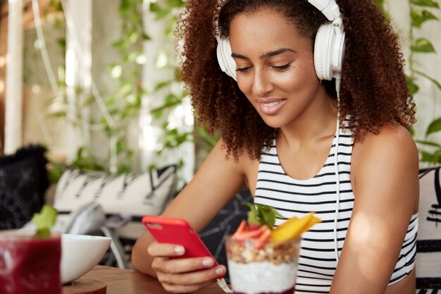 Dark skinned mixed race female in stylish headphones downloads audio book on mobile phone, spends free time at cafe, listens electronic music. Happy woman chooses favourite song in playlist.