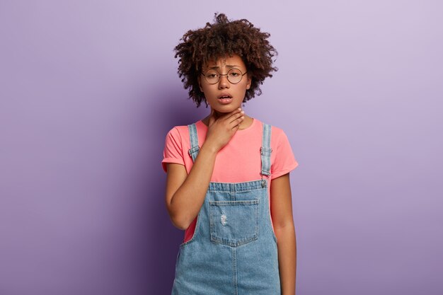 Dark skinned unhappy woman holds inflamed throat, feels pain while swallows, has cough, dressed in stylish clothes, spectacles isolated over purple wall. Bad symptoms. Problems with health