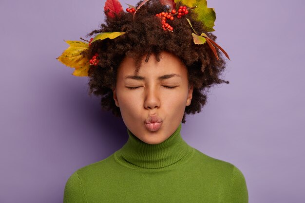 dark skinned woman has rounded lips, closes eyes, decorated head with autumn fallen leaves, dressed in casual wear