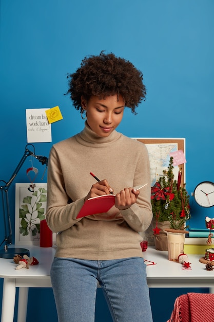 Free photo dark skinned woman stands indoor, wears turtleneck and jeans, writes down notes in diary with pencil