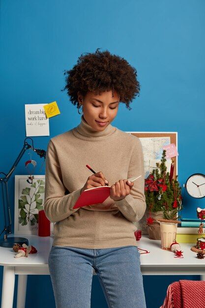 dark skinned woman stands indoor, wears turtleneck and jeans, writes down notes in diary with pencil