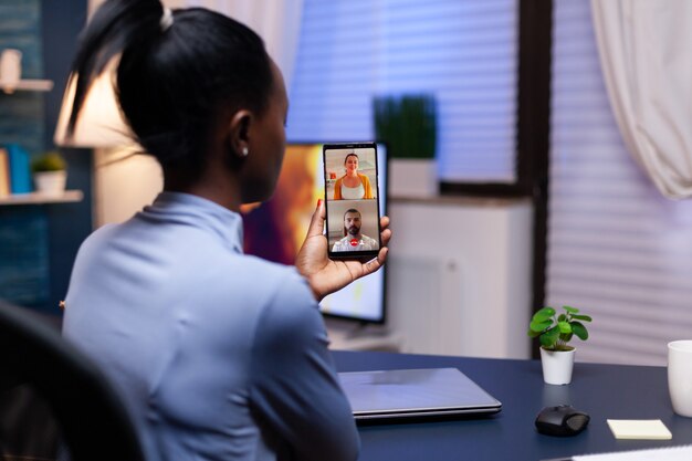 スマートフォンでのビデオ会議の過程で深夜に同僚とプロジェクトについて話している浅黒い肌の女性。仕事のために残業をしている最新のテクノロジーネットワークワイヤレスを使用している忙しい従業員。