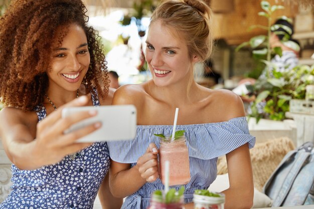 Dark skinned young female and friend has positive expression, make selfie, drink smoothie, spend recreation time at cafeteria. People, recreation time and lifestyle concept
