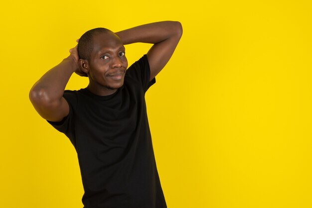 Dark-skinned young man standing his hands behind his head on yellow wall