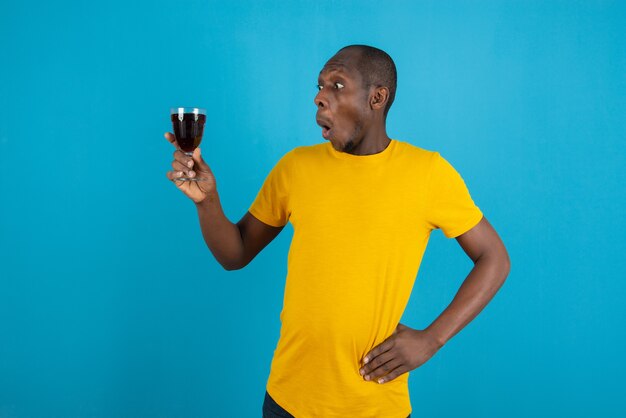 Dark-skinned young man in yellow shirt holding glass of wine on blue wall