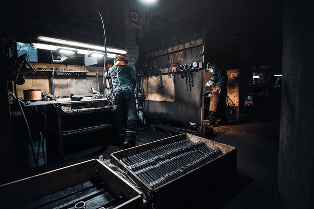 В темной мастерской с металлом работает опытный рабочий в защитной форме.