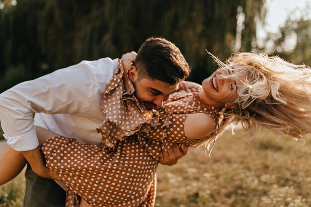 Darkhaired husband holding his beautiful wife in his arms laughing from tickling Portrait of couple fooling around in park in spring