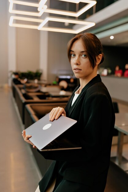 Темноволосая женщина в наушниках говорит по видеосвязи и держит ноутбук во время работы в современном офисе Женщина позирует перед камерой в офисе
