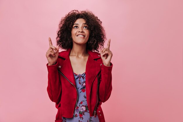 Darkskinned woman in red jacket shows fingers up on pink background Curly lady in floral dress smiling on isolated backdrop