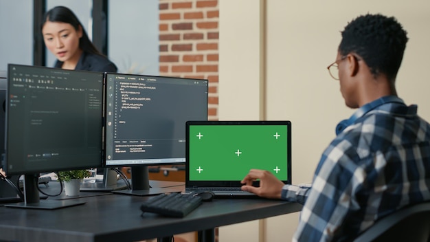 Free photo database designer writing code using laptop with green screen chroma key mockup sitting at desk in busy developers agency. programer looking at multiple screens with programming language.