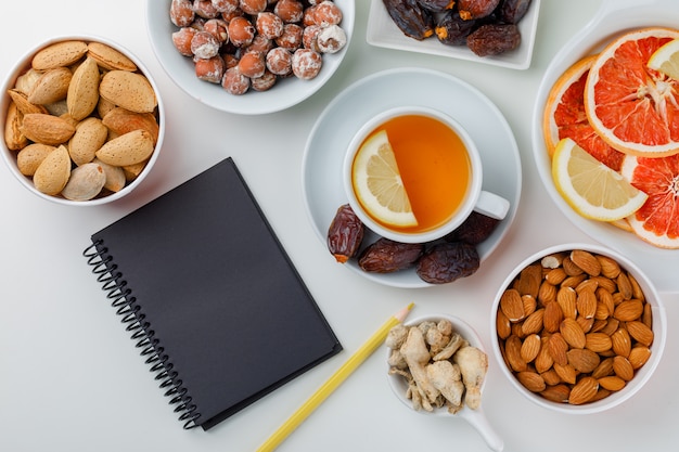 Dates, almonds, nuts in white plates with lemony tea, ginger, citrus fruits, pencil and notebook flat lay on a white table