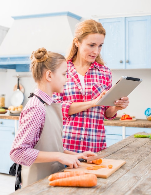 Daughter cutting the carrot looking at her mother using digital tablet