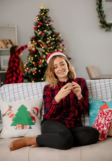 Дочь украшает елку, а мать в шляпе санта-клауса сидит на диване и наслаждается Рождеством дома
