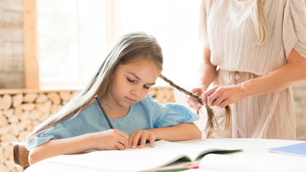 Daughter doing her homework at home while mother braids her hair