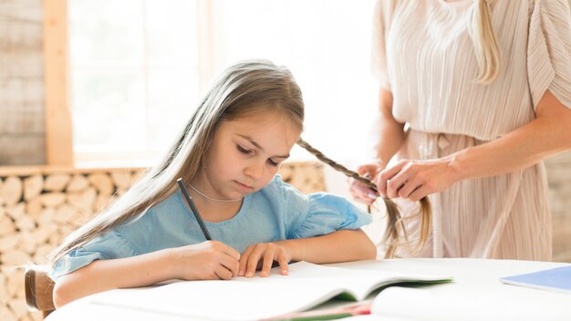 Daughter doing her homework at home while mother braids her hair