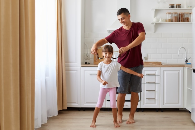 Free photo daughter and father having fun and dancing in the kitchen, people wearing casual clothing, man raising small girl pigtails, happy family spending time together at home.