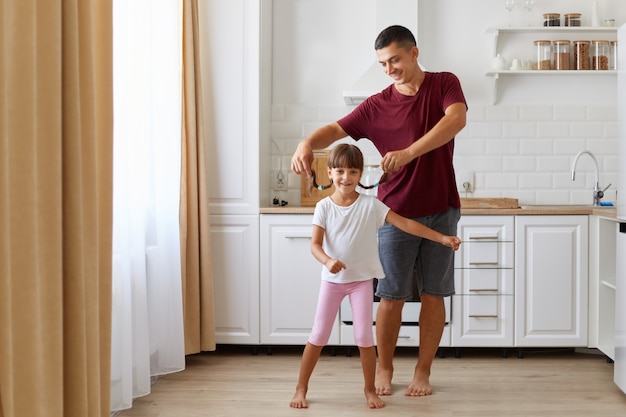 Figlia e padre che si divertono e ballano in cucina, persone che indossano abiti casual, uomo che alza le trecce da bambina, famiglia felice che trascorre del tempo insieme a casa.