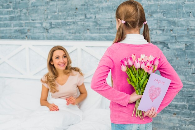 Daughter holding greeting card and tulips for mother in bed 