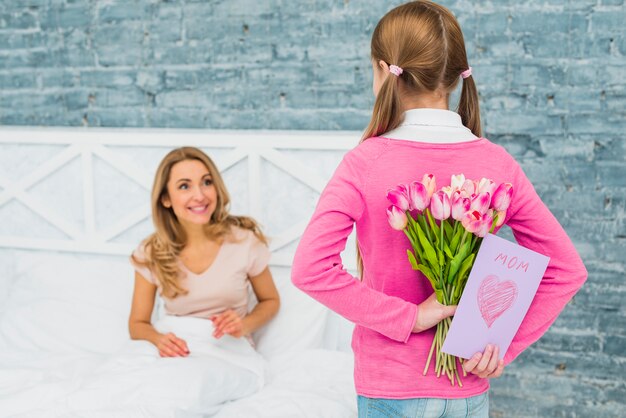 Дочь держит поздравительную открытку и тюльпаны для мамы в постели