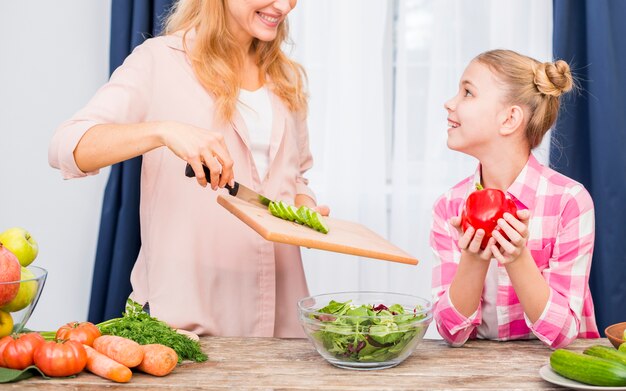 Daughter holding red bell pepper in hand looking at her mother preparing the salad