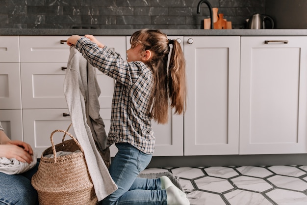 Daughter in jeans and shirt helps mom and pulls out dirty clothes from basket.