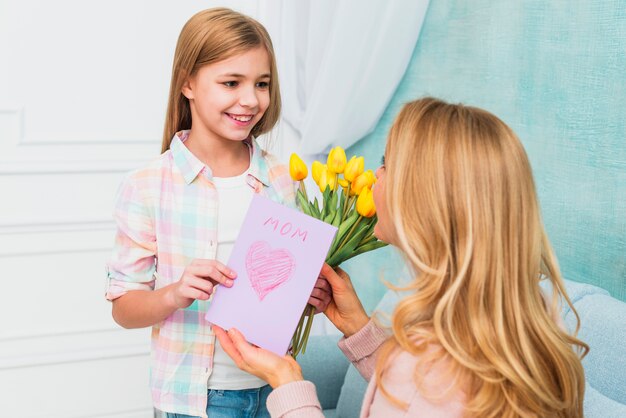 Daughter smiling and presenting flowers and Mother`s day postcard for mom