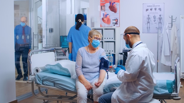 現代の民間クリニックや病院でのパンデミック時に、老婆の心臓関連の問題をチェックするDcotor。 COVID-19からの保護のためにマスクを着用している患者と医療用品。メディカルヘルスカ
