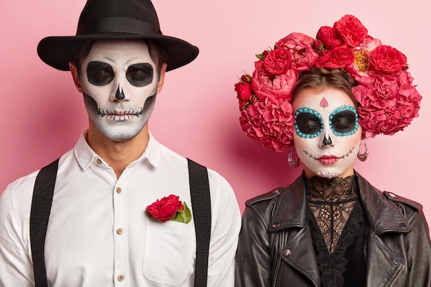 Dead spooky couple celebrate Halloween together, organize costume party, wear traditional mexican attire, vivid makeup, red flower wreath, pose in studio, stand shoulder to shoulder. Day of dead