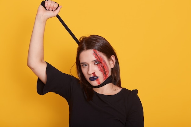 Free photo dead zombie female ready for halloween party, wearing black dress and scary makeup, choking herself with peace of cloth