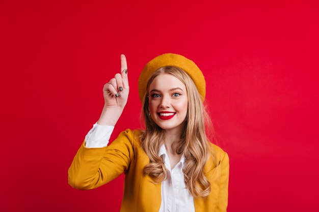 Debonair caucasian girl in beret pointing up with finger.  charming french lady gesturing on red wall.