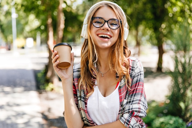 Foto gratuita signora caucasica debonair che esprime emozioni positive nel parco. foto all'aperto della donna splendida sorridente che beve caffè sulla natura.