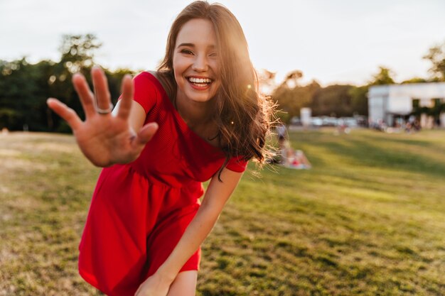 Debonair 백인 여자는 화창한 날에 카메라에 웃 고. 공원에 서있는 빨간 드레스에 쾌활 한 아름 다운 여자의 야외 사진.