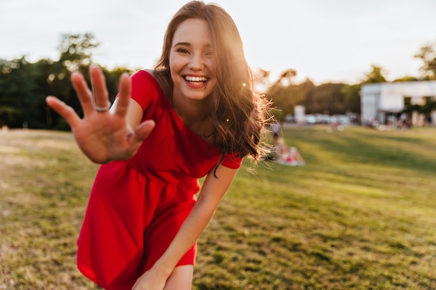 晴れた日にカメラに微笑んでいるDebonair白人女性。公園に立っている赤いドレスを着た陽気な美しい少女の屋外写真。