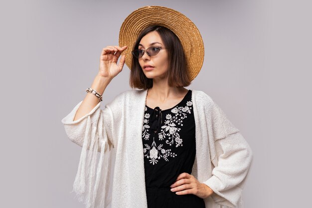 Жизнерадостная темноглазая девушка в соломенной шляпе и летнем наряде в стиле бохо позирует.